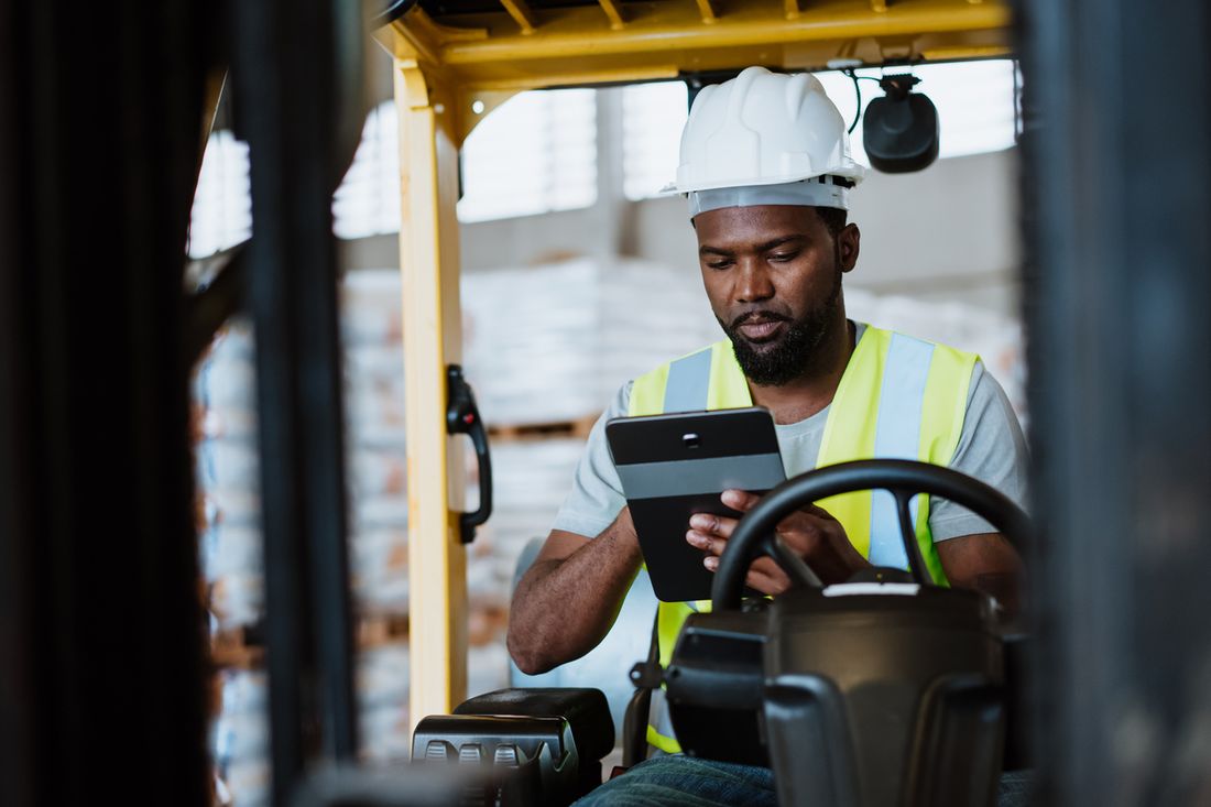 A forklift operator in a hard hat and safety vest using a tablet in a warehouse