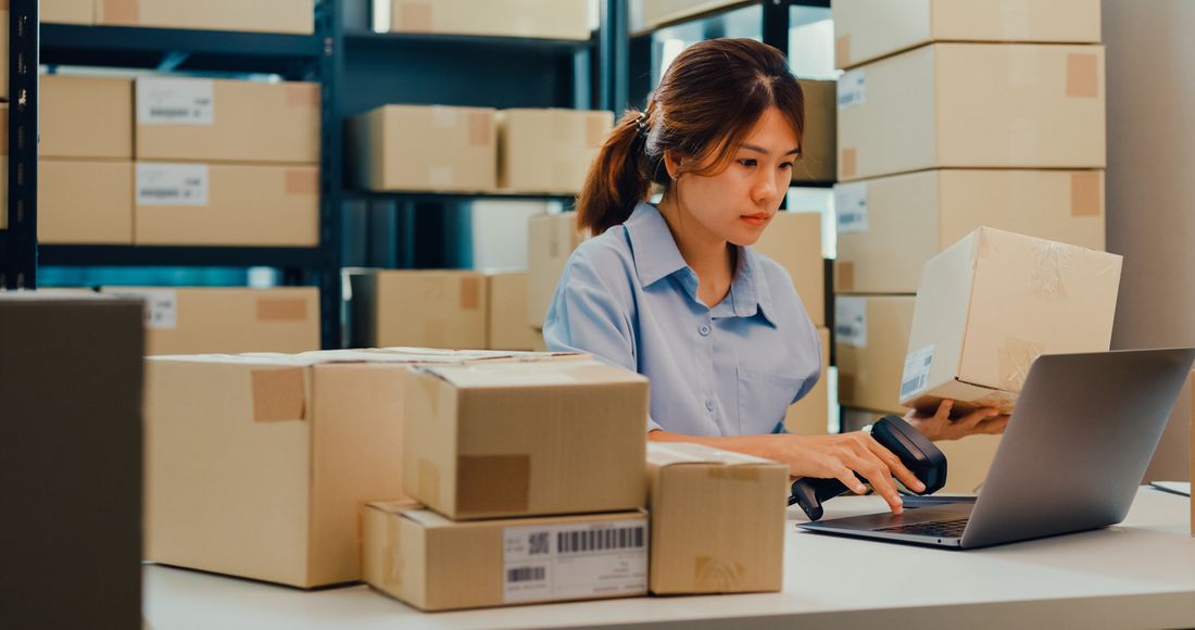 A worker scanning a package barcode at a laptop, surrounded by shipping boxes