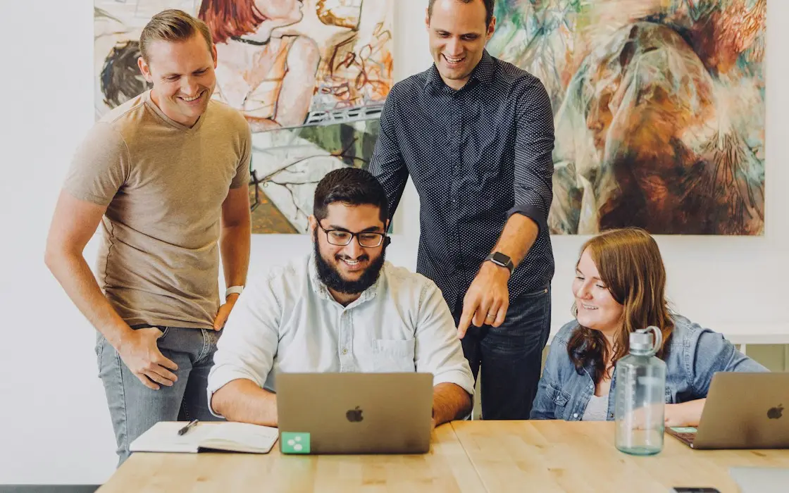 Four coworkers smiling around a laptop at a shared table