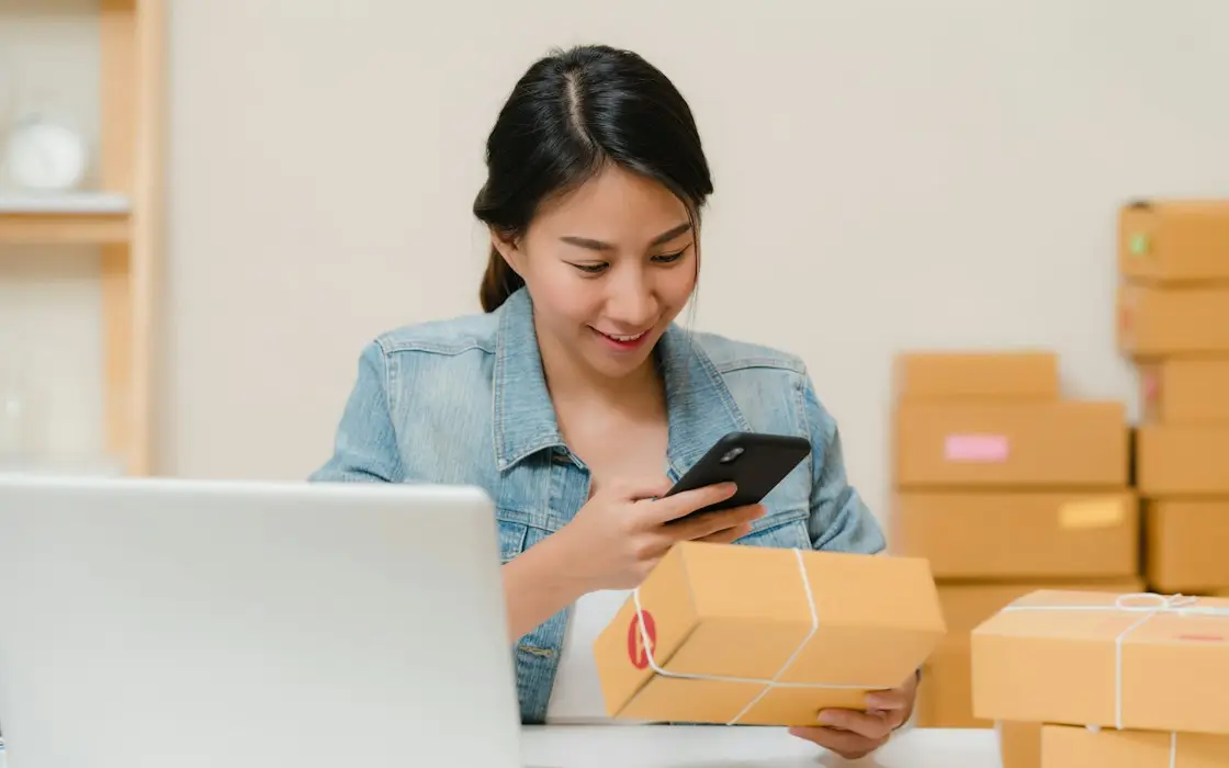 An online seller checking an order on her phone beside a laptop and shipping boxes